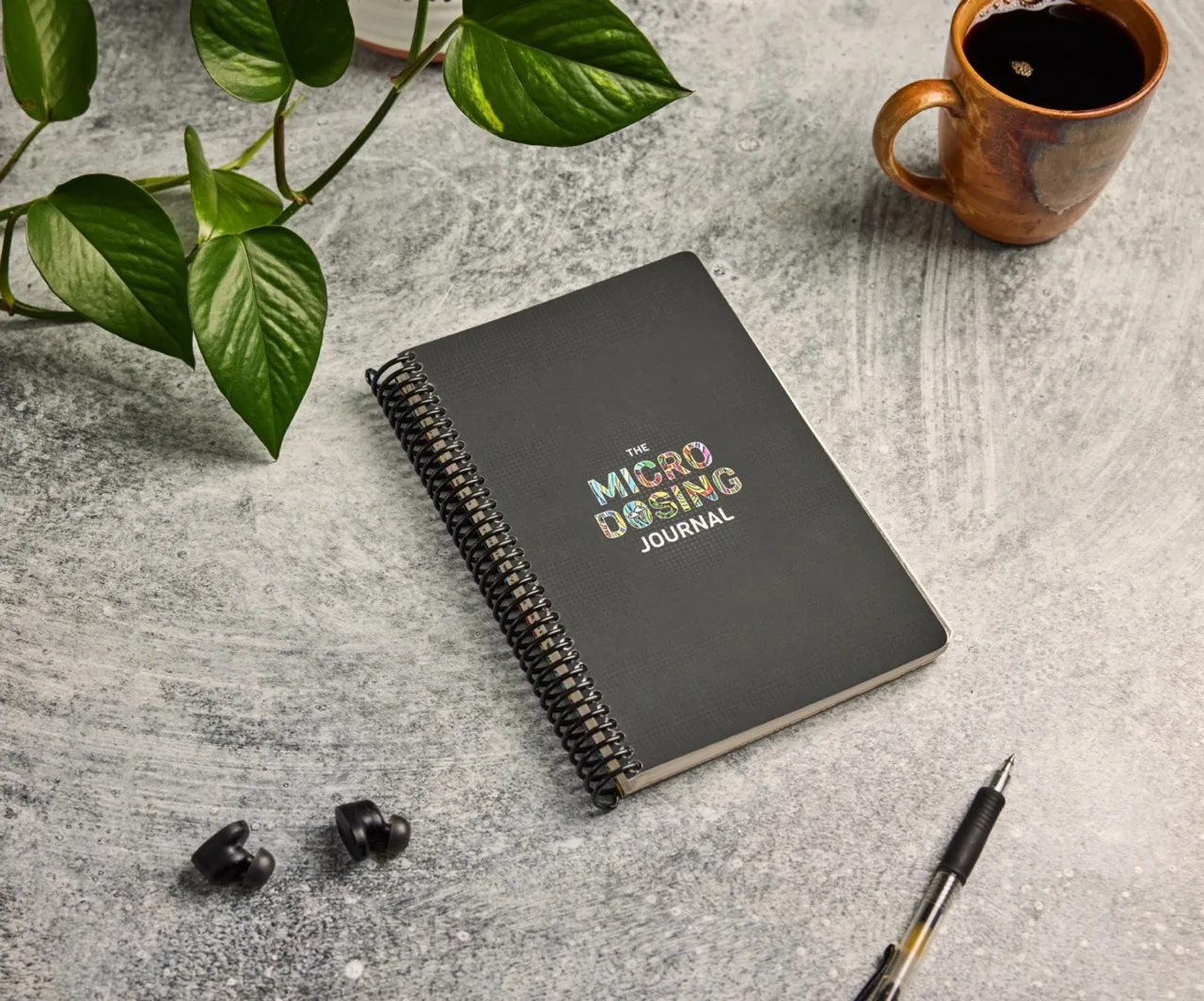 The Microdosing Journal cover on a gray desk beside a plant, coffee, pen, and earbuds