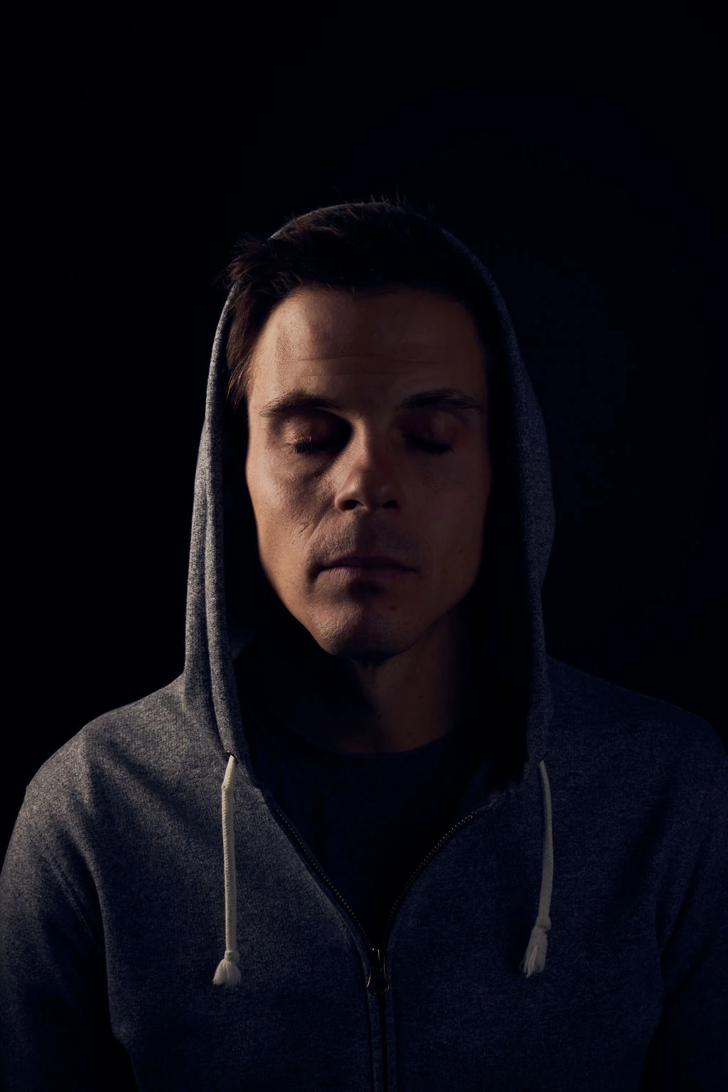 Seth Marcus with his eyes closed, wearing a gray hooded sweatshirt against a dark background.
