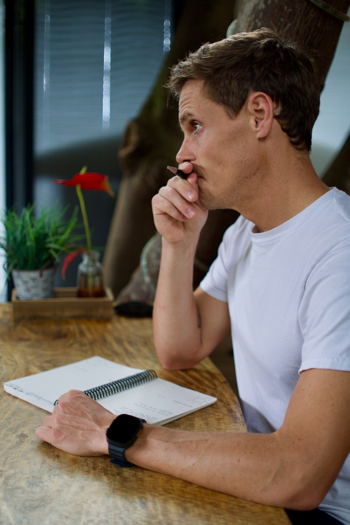 A man seated at a wooden table, holding a pen near his face with an open notebook in front of him.